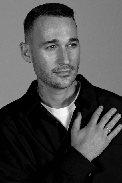Monochrome portrait of a man in a dark collared shirt, hand on his chest, and a serious facial expression against a light background.