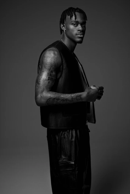 Monochromatic portrait of a Black male model in a vest and leather pants, posing in a photo studio setting, looking at the camera.
