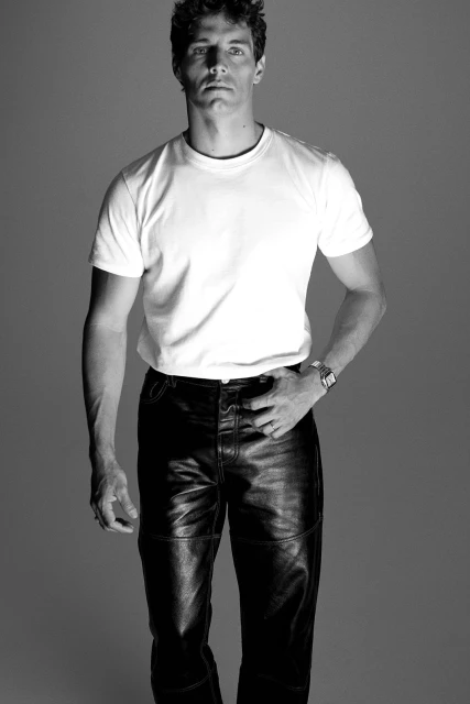 Monochrome portrait of a man in a white t-shirt and black leather pants posing in a photo studio under bright, even lighting.