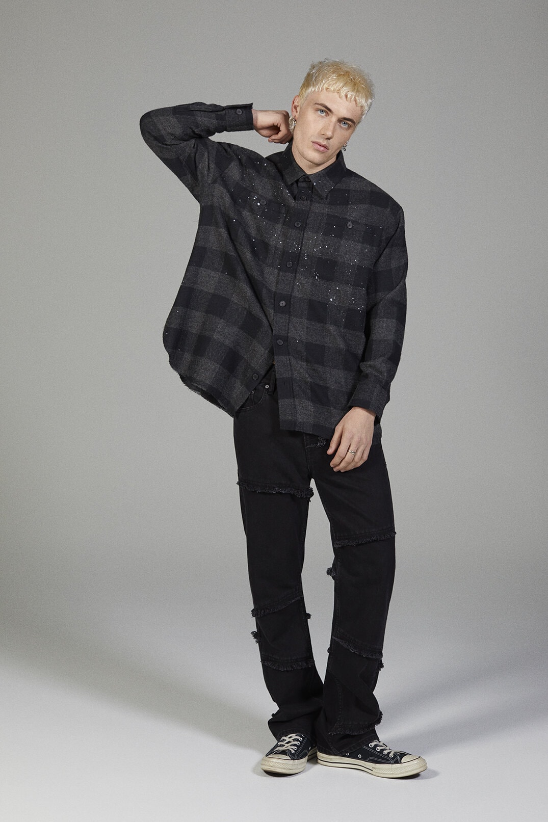 Man with blond hair poses in an open black and grey plaid shirt, black jeans, and black sneakers in a neutral studio setting.