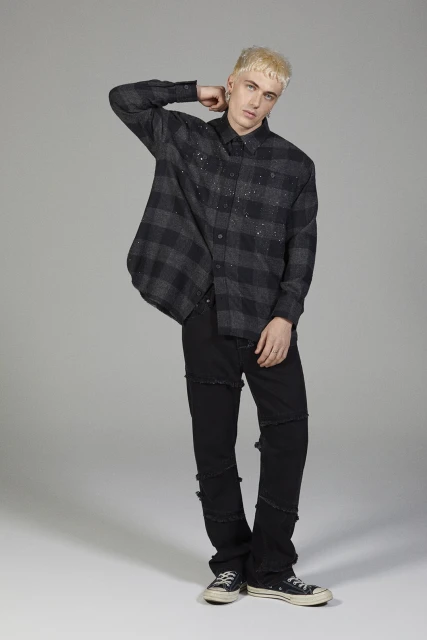 Man with blond hair poses in an open black and grey plaid shirt, black jeans, and black sneakers in a neutral studio setting.