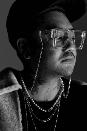 Monochrome portrait of a man wearing clear square glasses with a chain, layered pearl necklaces, and a dark cap against a grey background.