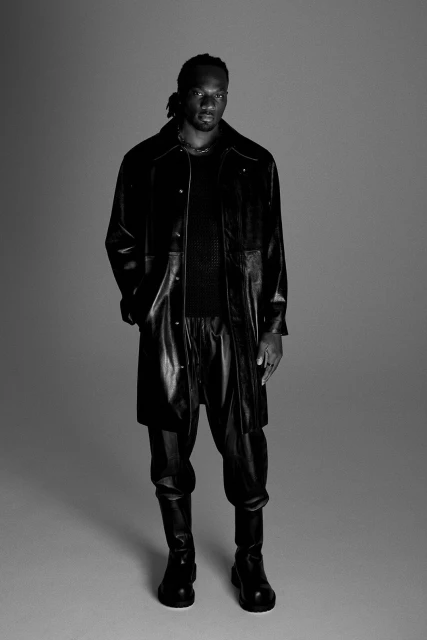 Black and white portrait of an African American man in a long, black leather coat with matching pants and boots in a photo studio.