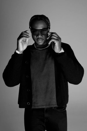 Portrait of an adult male with sunglasses, wearing a cardigan and jeans, posing indoors in a photo studio against a gray backdrop.
