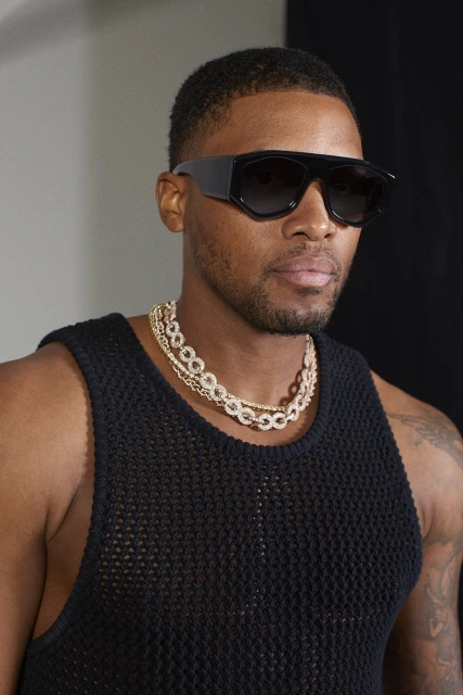 Close-up shot of an adult Black male wearing oversized sunglasses and a black tank top, styled with gold chain necklaces in a studio.