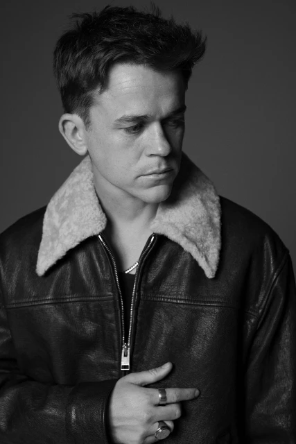 Black and white portrait of a man wearing a leather jacket with a shearling collar, looking down with a serious expression.