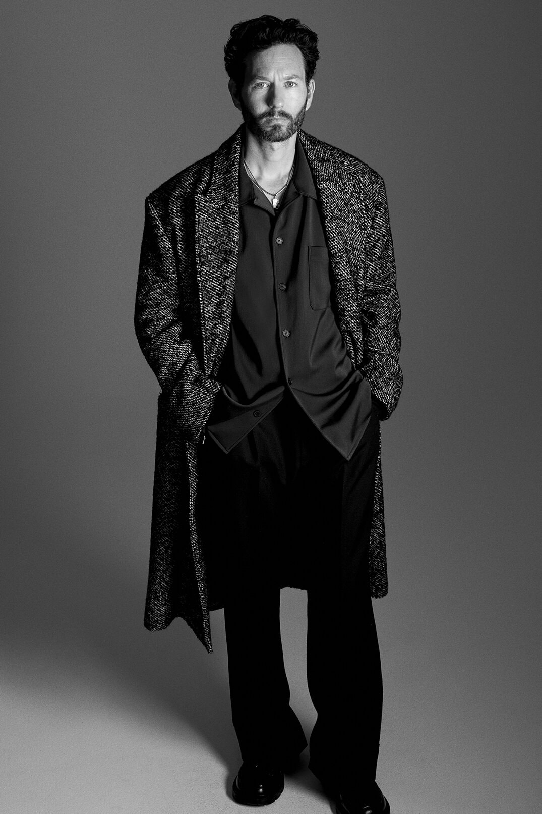 Monochrome portrait of a stylish man in a patterned overcoat, collared shirt, and dark trousers, standing in a neutral studio setting.