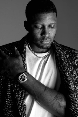 Close-up of a Black man with short hair wearing a textured blazer over a white t-shirt, silver necklace, and wrist watch against a grey wall.