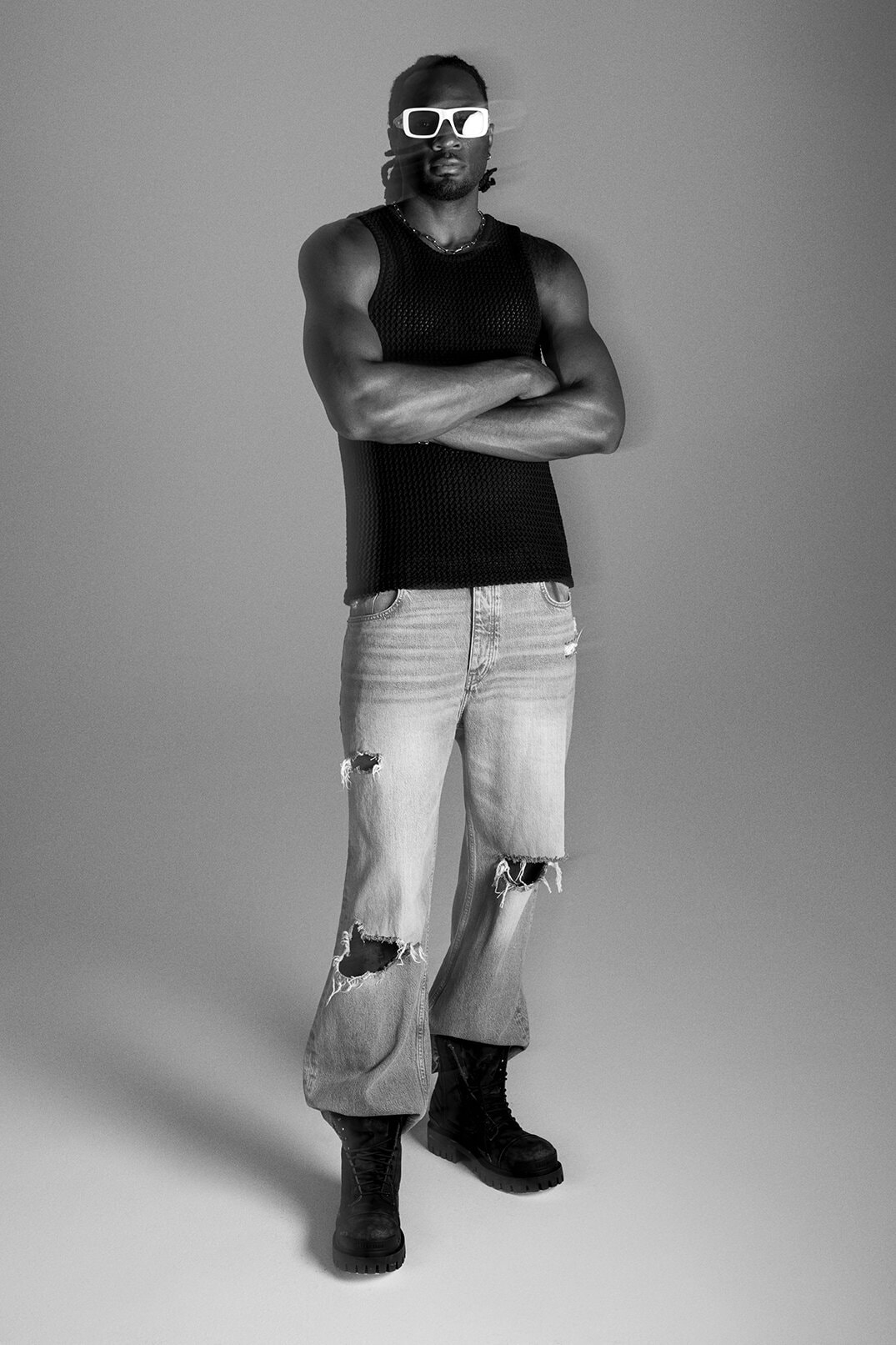 Man wearing sunglasses and black tank top standing against a grey studio backdrop for FRAME Modern Man campaign