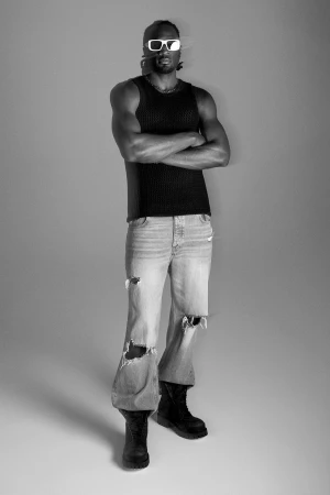 Man wearing sunglasses and black tank top standing against a grey studio backdrop for FRAME Modern Man campaign