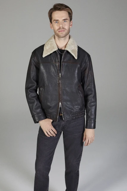 A man in a leather jacket with a fur collar stands in a photo studio in dark jeans posing for a portrait against a grey background.