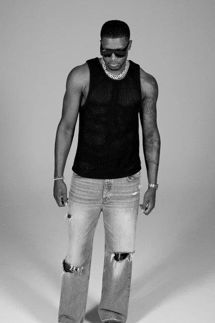 Monochrome portrait of an African American man in sunglasses, black tank top, and ripped jeans standing in a photo studio.