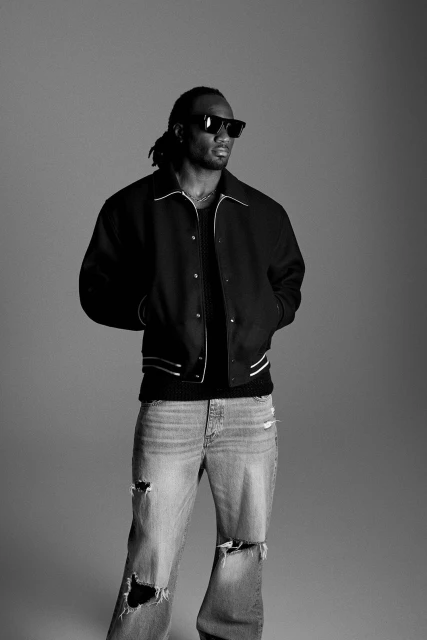 An adult male with dreadlocks wearing a dark letterman jacket, light-wash distressed denim jeans, and sunglasses posing in a studio.