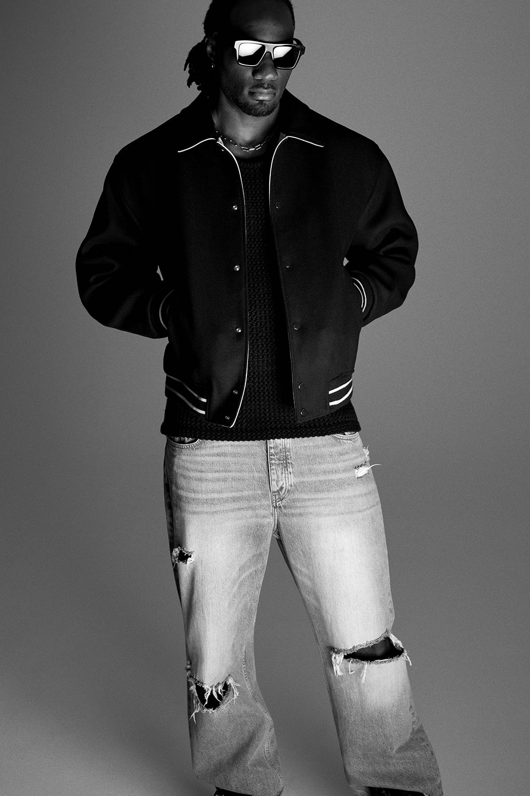 Black and white portrait of a man in a black varsity jacket and ripped jeans. He is also wearing sunglasses in an indoor studio.