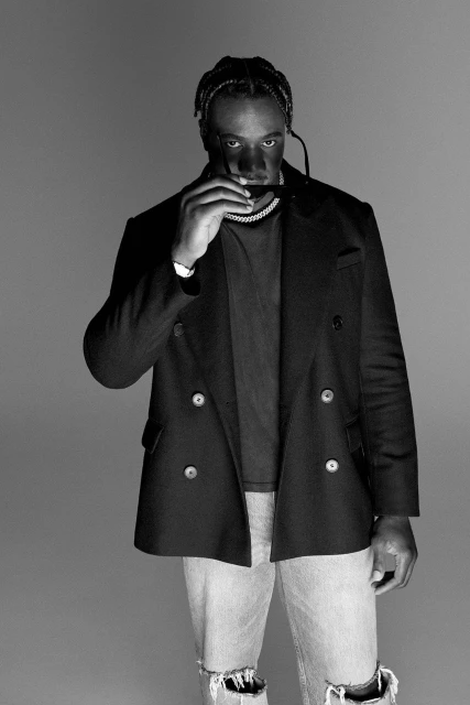 Black and white portrait of an adult male wearing a black double-breasted jacket and ripped jeans, holding sunglasses in front of his face.