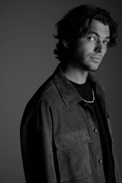 Black and white portrait of a young adult male in a buttoned overshirt looking at the camera, with a necklace, in a photo studio.