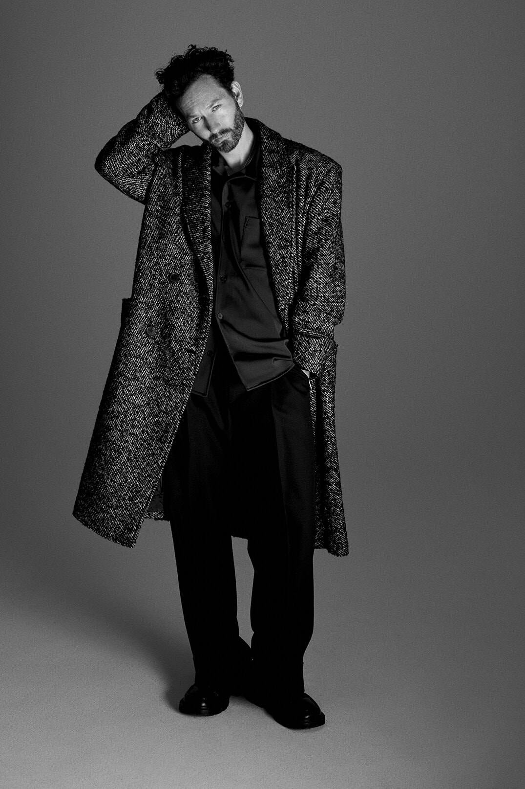A man standing in a textured tweed overcoat and dark trousers with his hand behind his head against a grey studio backdrop.