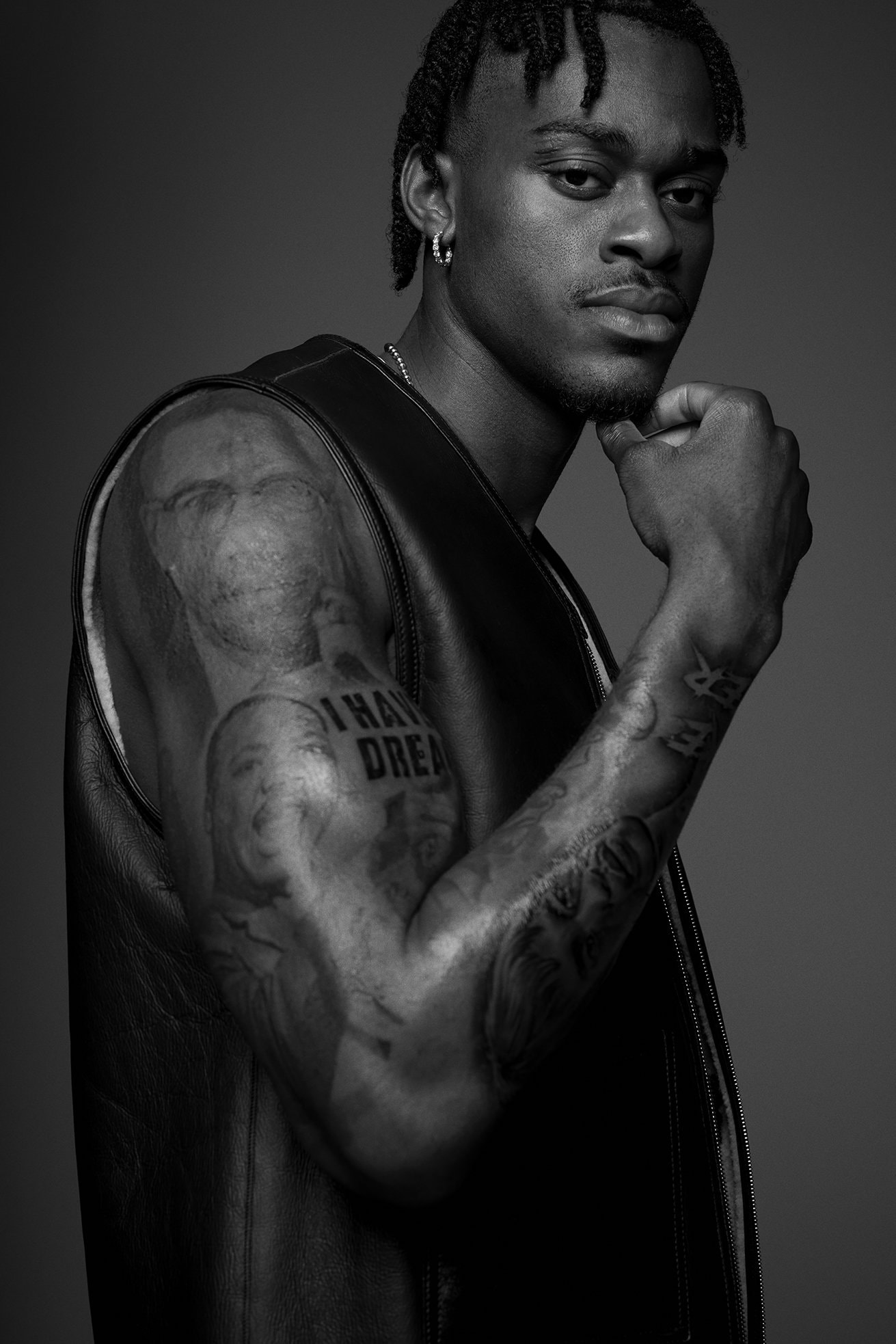 Black and white portrait of a young adult man with braided hair and tattoos, wearing a vest and posing in a photo studio.