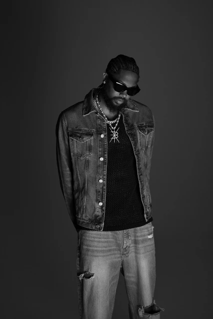 Portrait of a Black man with braids wearing a denim jacket, black top, distressed jeans, sunglasses, and jewelry against a grey backdrop.