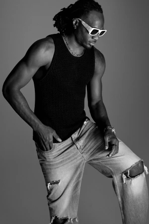 Black man wearing sunglasses, a black tank top, and distressed jeans against a grey studio backdrop.