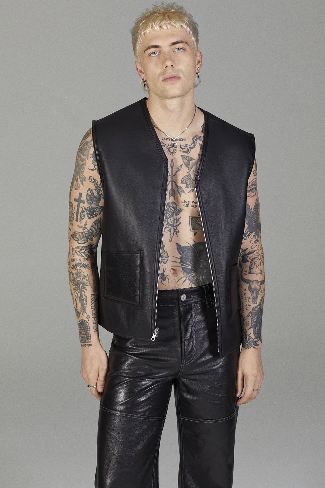 Young adult male with tattoos poses in a black leather vest and black leather pants, indoor studio environment with neutral backdrop.