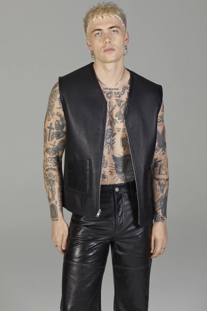 Young adult male with tattoos poses in a black leather vest and black leather pants, indoor studio environment with neutral backdrop.