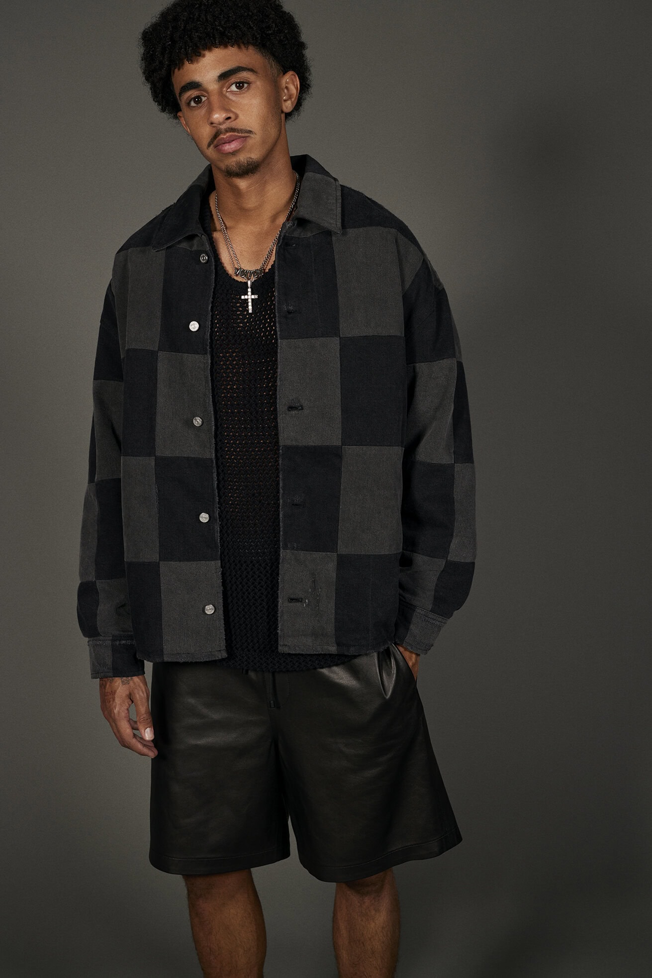 A young adult male poses in a checkered jacket and black shorts, gazing seriously at the camera in a fashion-forward indoor portrait.