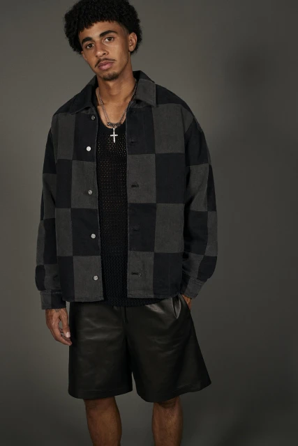 A young adult male poses in a checkered jacket and black shorts, gazing seriously at the camera in a fashion-forward indoor portrait.