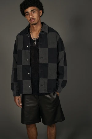 A young adult male poses in a checkered jacket and black shorts, gazing seriously at the camera in a fashion-forward indoor portrait.
