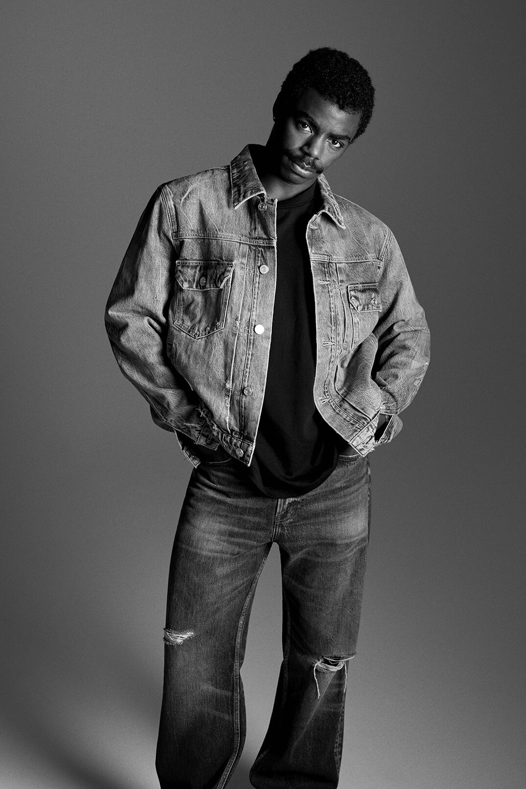 Black man in denim jacket and jeans poses in a studio with a neutral gray background, creating a stylish and relaxed fashion portrait.
