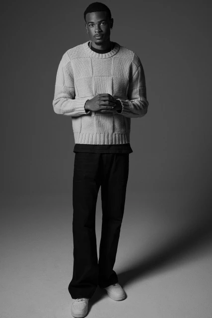 Studio shot of a Black male model in a beige knit sweater and black pants, hands clasped, looking directly at the camera. Monochrome aesthetic.