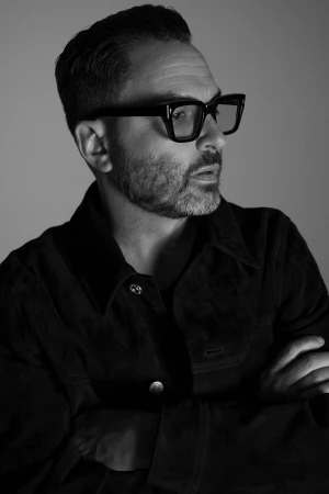 Monochrome portrait of a man wearing thick-rimmed glasses and a dark jacket with arms crossed against a grey backdrop