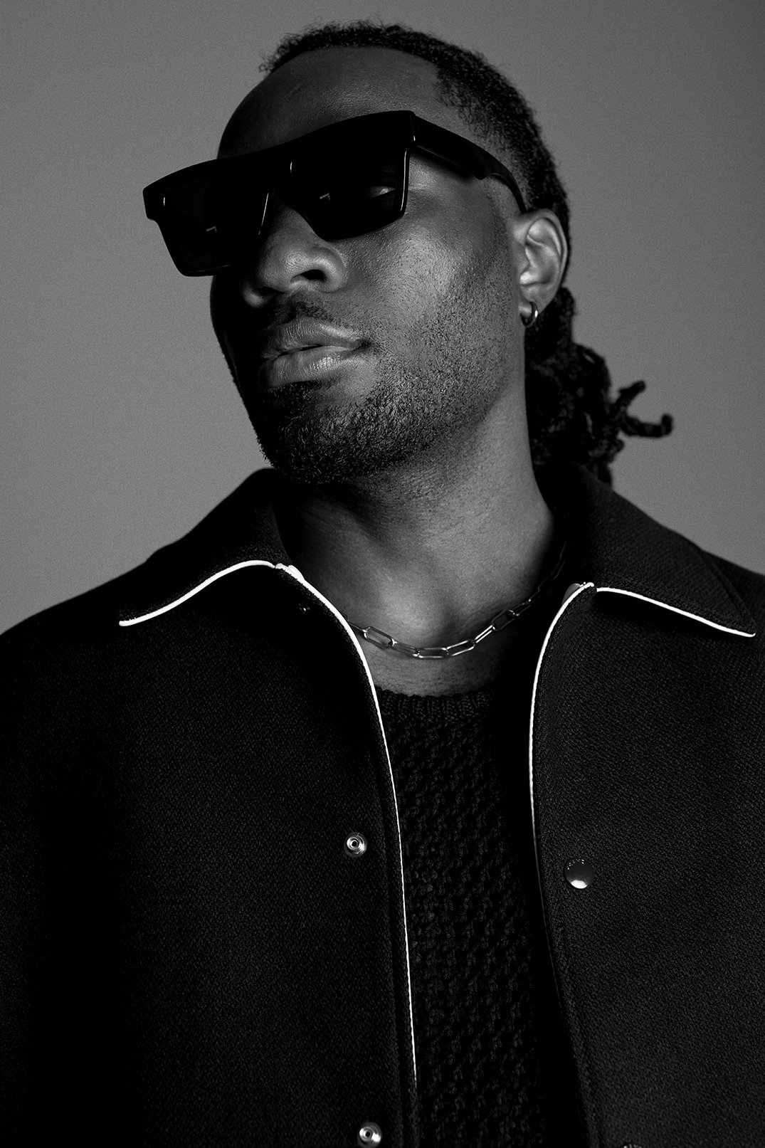 Monochrome portrait of a Black man wearing dark sunglasses and a black jacket with white piping against a grey studio backdrop.
