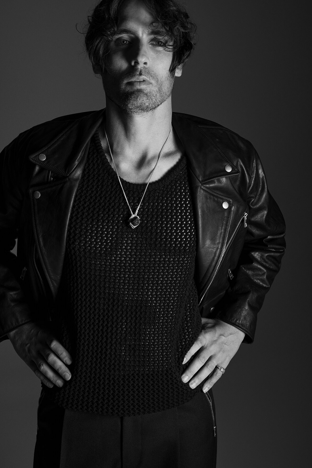 Black and white portrait of an adult male in a leather jacket and knitted sweater, posing in a studio with hands on his hips.