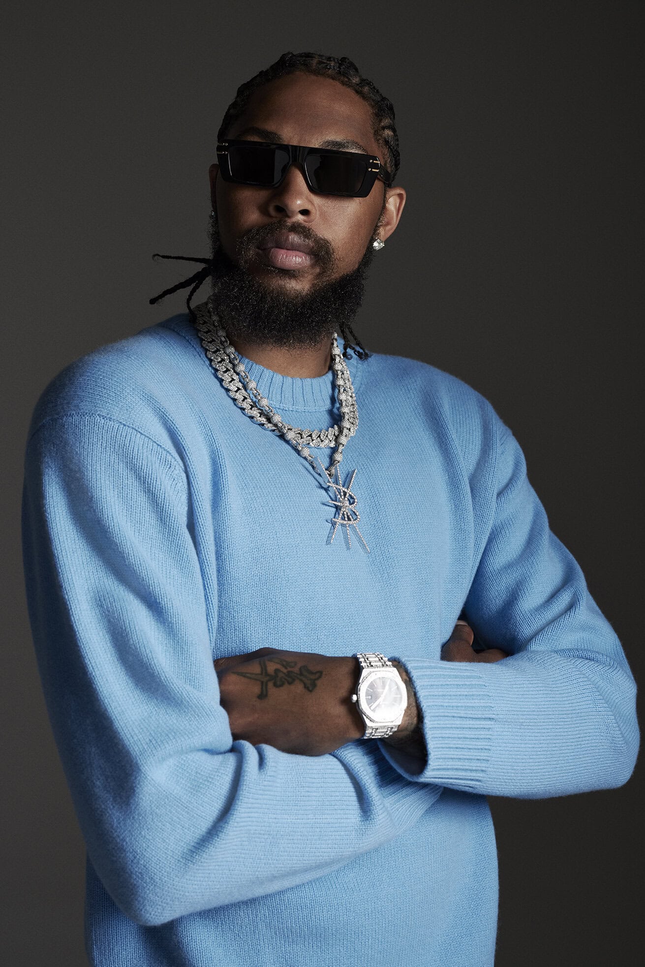 Portrait of a Black male adult in a blue sweater, wearing sunglasses and jewelry against a grey backdrop in a studio setting.