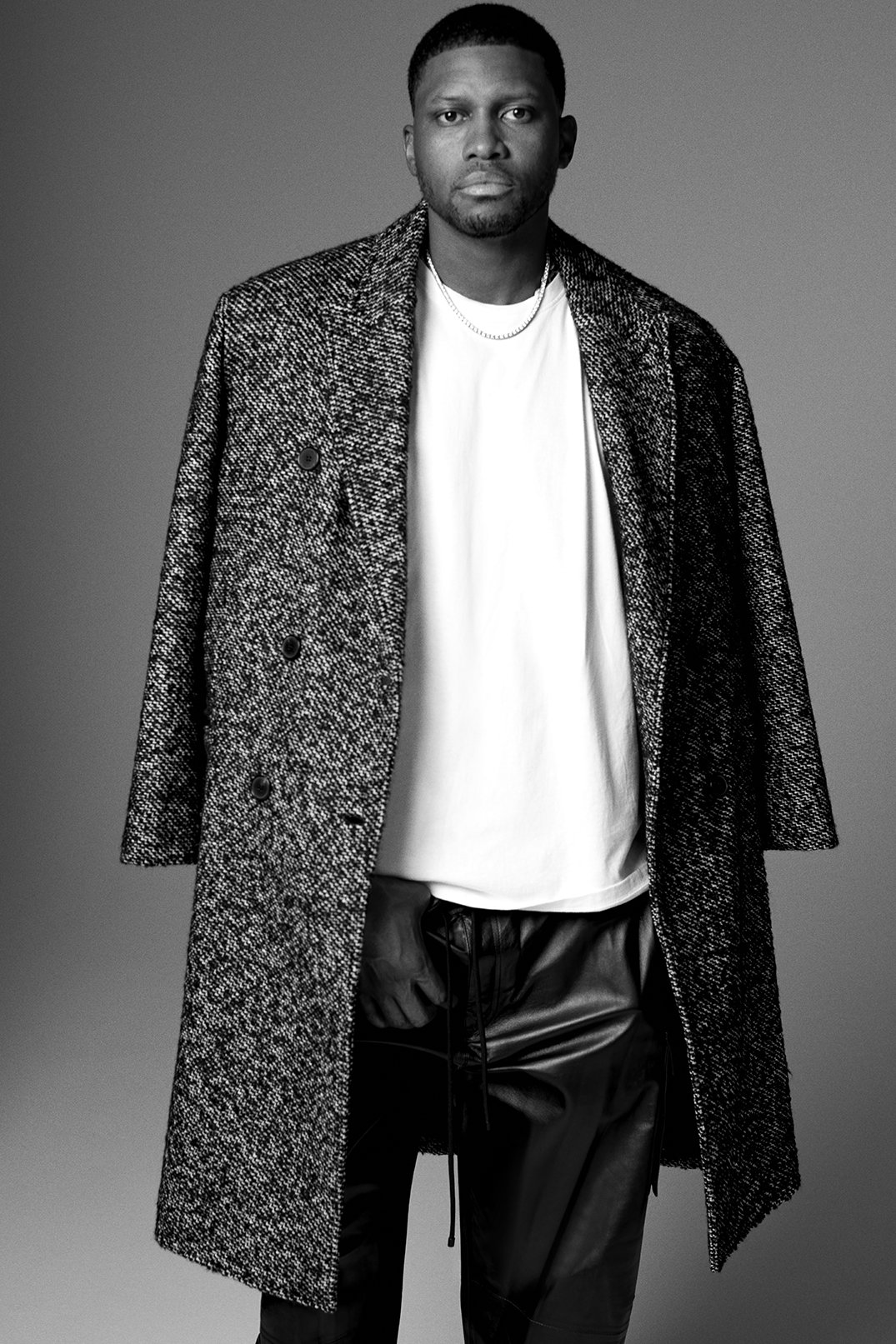 Black and white portrait of a Black man wearing a long coat and leather pants against a grey background in a studio setting.