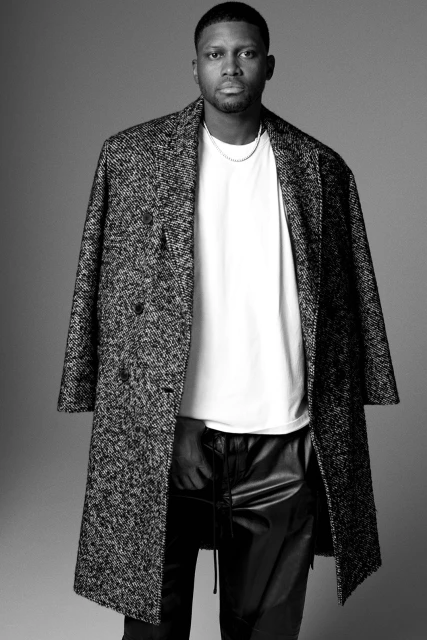 Black and white portrait of a Black man wearing a long coat and leather pants against a grey background in a studio setting.