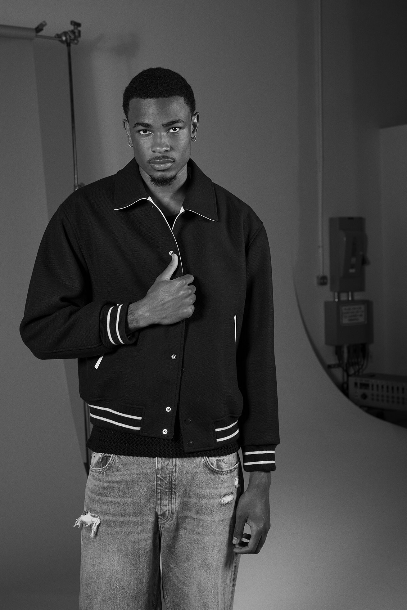 Monochrome portrait of a stylish young man in a varsity jacket and ripped jeans posing in a photo studio, bathed in soft lighting.
