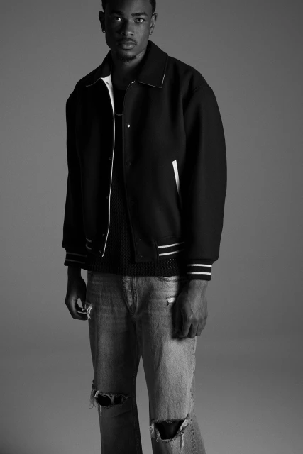 Full-length portrait of a young adult male in a black and white varsity jacket with distressed jeans, set against a gray background.