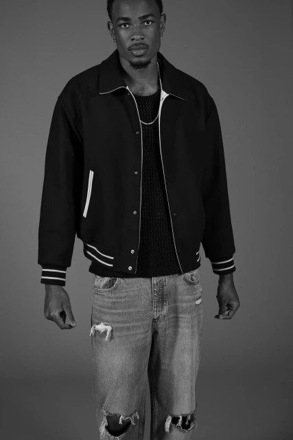 Black and white portrait of a Black man in a varsity jacket and distressed jeans standing in a photo studio with a neutral grey backdrop.