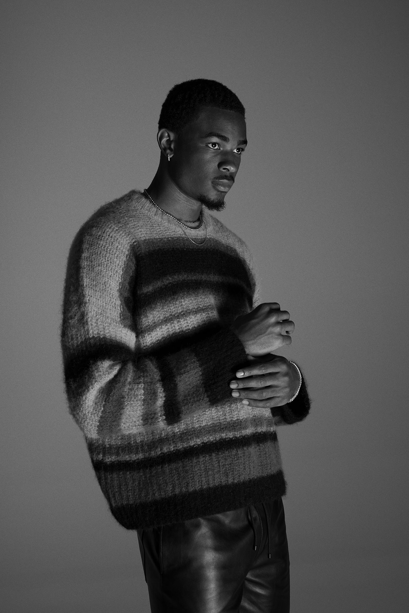 Black man in a striped sweater and leather pants posing in a grey studio. Fashionable outfit for a photoshoot in a studio environment.