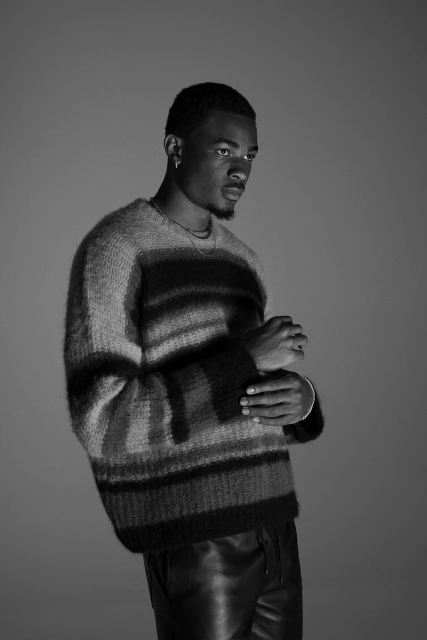 Black man in a striped sweater and leather pants posing in a grey studio. Fashionable outfit for a photoshoot in a studio environment.