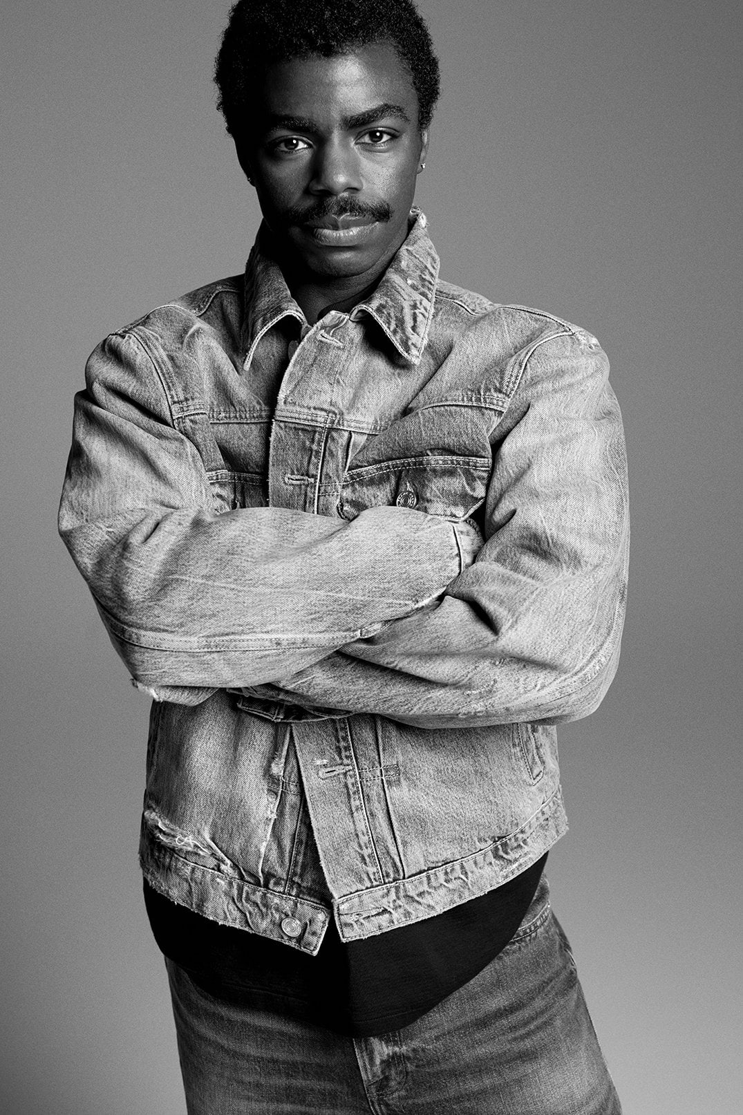 Black and white portrait of a Black male model with arms crossed, wearing a denim jacket and jeans against a grey background.