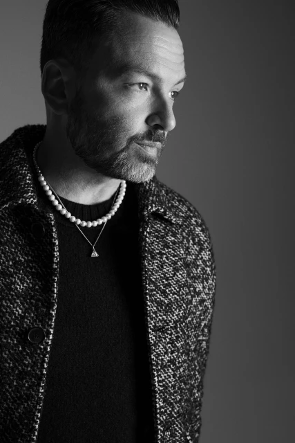 Portrait of a stylish adult male in black and white, wearing a pearl necklace and a tweed jacket, in a studio setting.