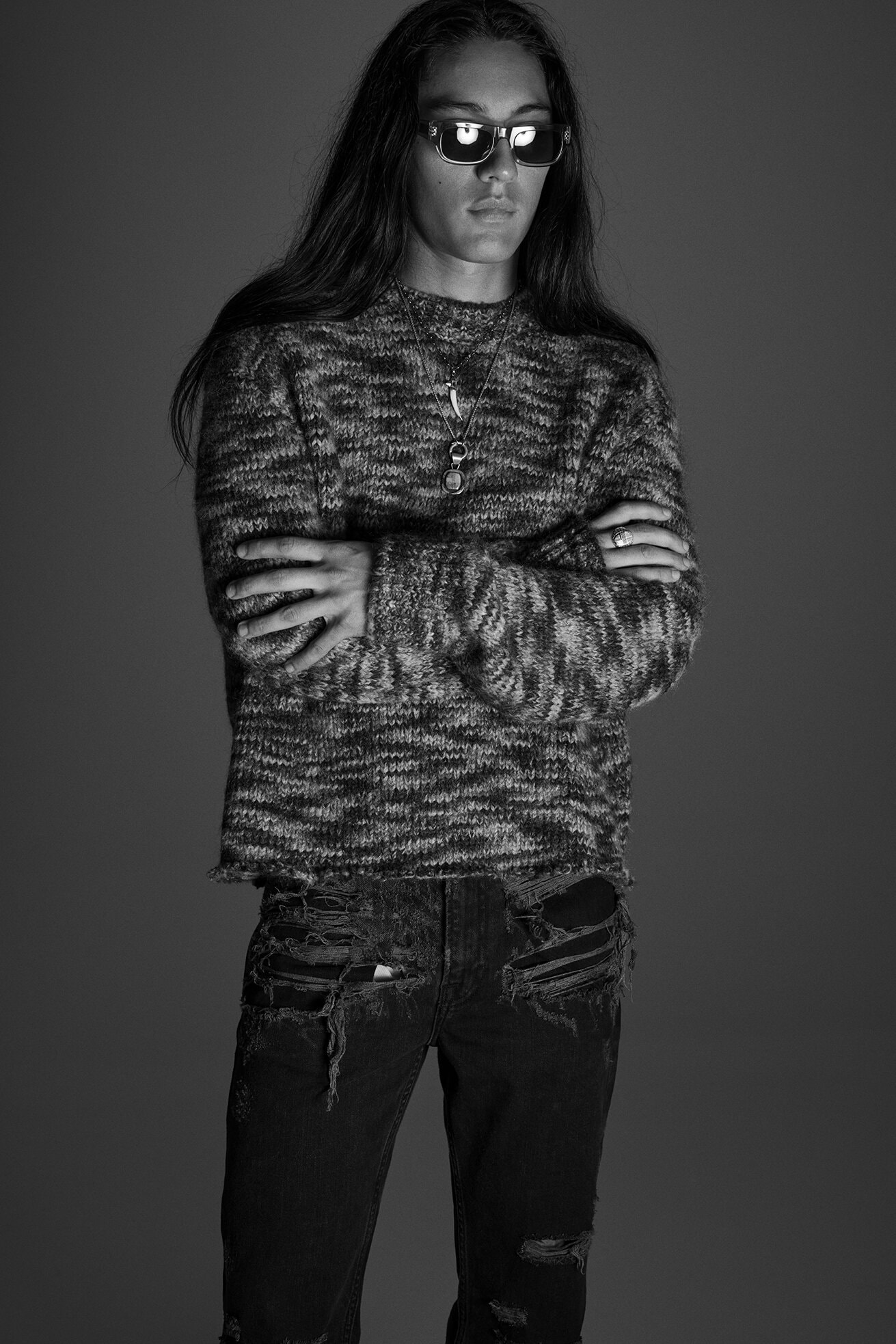Black and white portrait of a long-haired man in sunglasses, wearing a sweater and ripped jeans, against a grey backdrop.