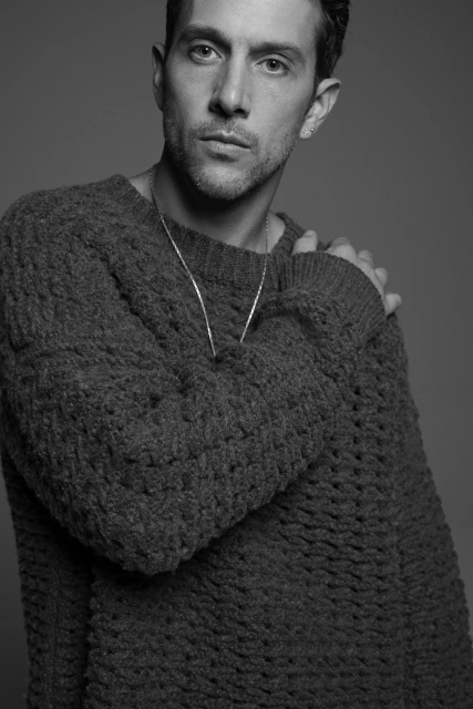 Black and white portrait of a man wearing a textured sweater and a necklace in a photo studio with a grey backdrop.