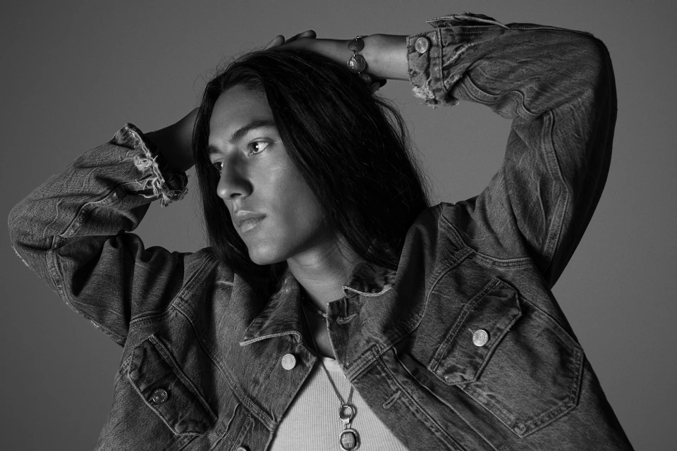 Monochrome portrait of a young adult with long dark hair, wearing a denim jacket and jewelry, posing against a grey studio backdrop.