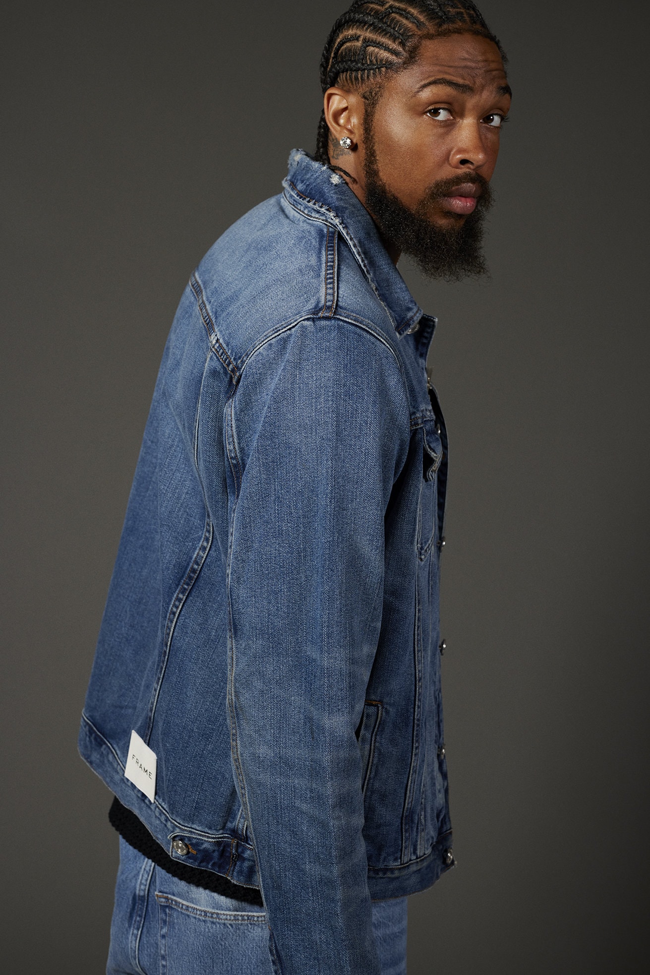Adult male with cornrow braids and beard wearing a blue denim jacket, looking at the camera in a studio portrait against gray.