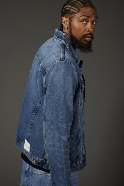 Adult male with cornrow braids and beard wearing a blue denim jacket, looking at the camera in a studio portrait against gray.