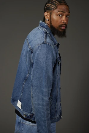 Adult male with cornrow braids and beard wearing a blue denim jacket, looking at the camera in a studio portrait against gray.